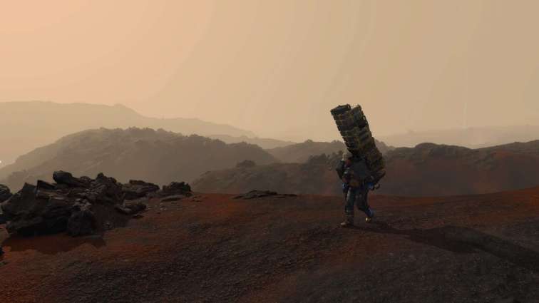 Sam trudges across a reddish brown landscape, a tall stack of cargo strapped to his back.